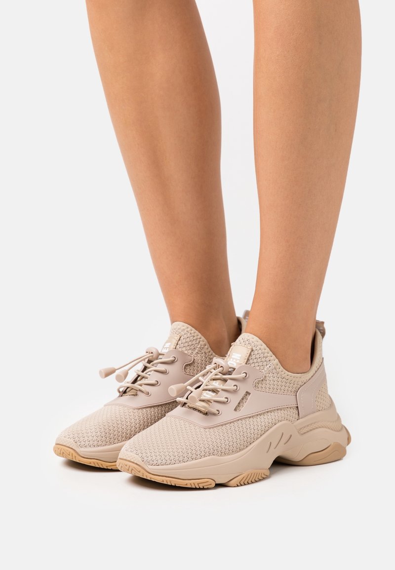 Pieds portant des baskets beiges en tricot avec des semelles épaisses en caoutchouc et des lacets élastiques sans nœud sur fond blanc.