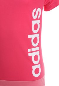 Rosa tränings-t-shirt i mjukt material, med en stor vit "adidas" logotyp vertikalt längs vänster sida. Kortsleeves och åtsittande design.