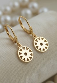 Orecchini circolari in oro con design a stella ritagliata, appesi a ganci a leva, esposti su un tessuto beige texture.