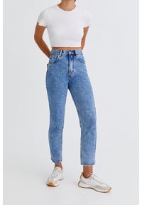 Jean taille haute bleu clair avec un délavage légèrement usé, silhouette ajustée, cinq poches et passants de ceinture, associé à un top blanc court.