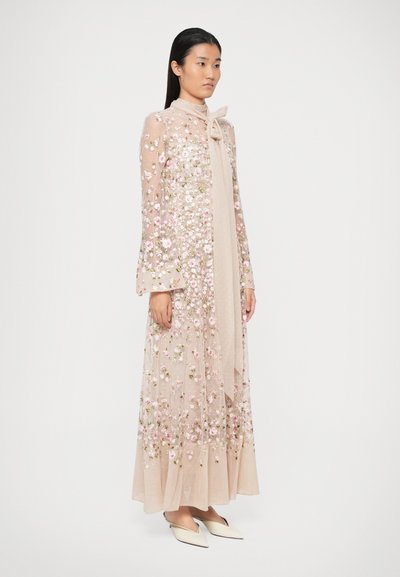 Γυναίκα που φοράει ένα μακρύ ημιδιαφανές μπεζ φόρεμα με λουλουδάτη κεντητική, χυτές μα�νίκες και ένα κομψό φουλάρι στον λαιμό, συνδυασμένο με λευκές μυτερές γόβες.