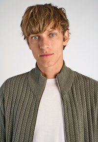 Light green knitted bomber jacket with a ribbed texture and a high collar, paired with a plain white t-shirt beneath.