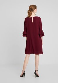 Femme portant une robe bordeaux mi-longue avec des manches trois-quarts à volants et des chaussures noires à talons hauts avec bride à la cheville, debout devant un fond uni.