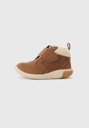 Bota de tornozelo em camurça castanha com sola em borracha bege e fita de velcro. Apresenta um colarinho forrado de pêlo e detalhes texturizados na parte superior.