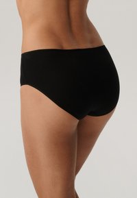 Kobieca tors i górna część ud w czarnych majtkach z wysokim stanem, pokazane z tyłu na tle jasnym.