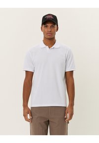 Chemise polo blanche en mélange de coton avec un col classique et des manches courtes, portée avec un pantalon cargo marron ; branding discret sur la poitrine.