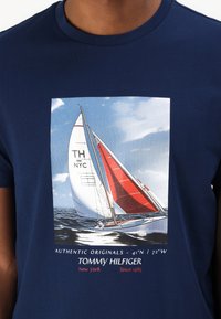 Mørkeblå bomull t-skjorte med et seilbåt grafikk med røde seil, hvit skrog, og tekst som detaljer "AUTENTISK ORIGINALER" og "TOMMY HILFIGER."