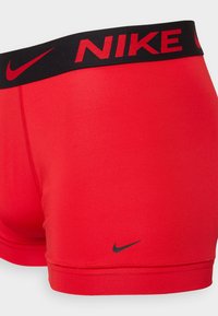 Shorts atletici roșii cu talie neagră, având inscripția "NIKE" în litere îngroșate. Textură netedă, design ajustat și logo subtil pe margine.
