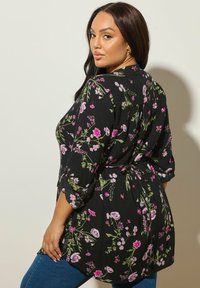 Blusa nera a fiori con motivi rosa e verdi, maniche lunghe, vita regolabile con nastro e tessuto testurizzato. Indossata sopra jeans blu.