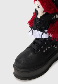 Stivale platform nero in pelle sintetica testurizzata, con occhielli in argento, lacci e un accessorio a forma di bambola con capelli rossi e un vestito nero.