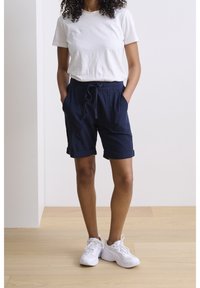 Pantaloni corti blu navy con una vestibilità rilassata, vita con cordino, orli risvoltati e tasche laterali, abbinati a una maglietta bianca semplice e sneakers bianche.