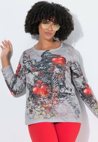 Frau mit lockigem Haar, die gelb getönte Pilotenbrille, ein graues Langarmoberteil mit roten Herz- und Blumenmotiven sowie leuchtend roten Hosen trägt.