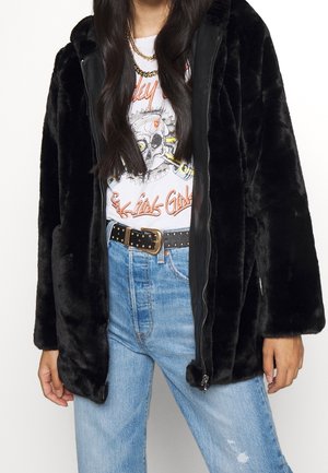 Woman wearing black faux fur coat over graphic skull t-shirt, light blue high-waisted jeans, studded black belt, and gold chain necklace.