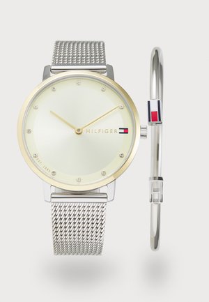 Montre avec bracelet en maille argenté, lunette en ton or, cadran vert clair et accents en clou. Comprend le logo de la marque et un petit bracelet rigide avec des accents de couleur.