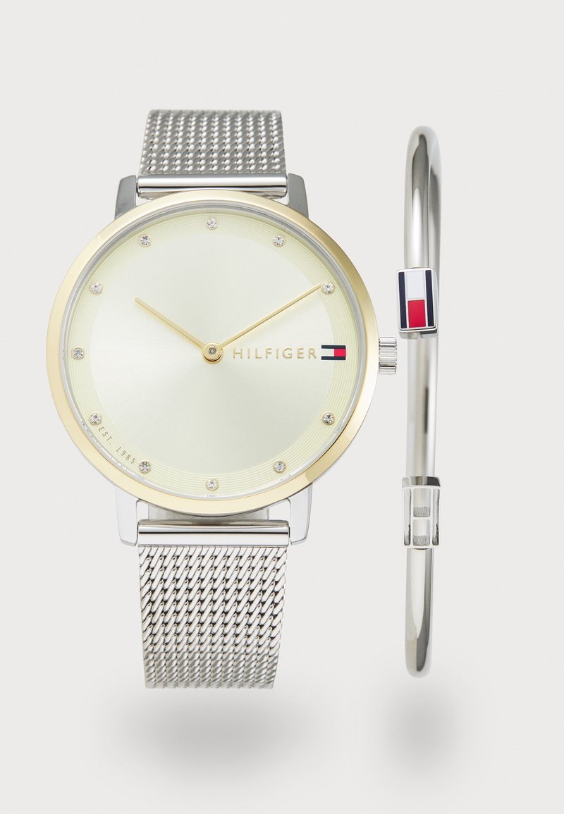 Reloj con pulsera de malla plateada, bisel en tono dorado, esfera verde clara y acentos de tachuelas. Presenta el logotipo de la marca y una pulsera más pequeña con acentos de color.