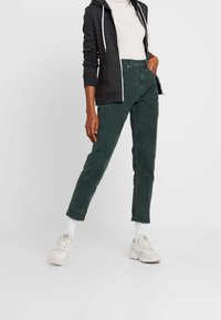 Osoba stojící před jednoduchým pozadím, oblečená v tmavě zelených kalhotách s vysokým pasem, bílých teniskách, bílých ponožkách, bílém roláku a černé mikině na zip.