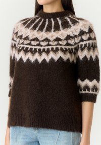 Femme portant un pull en laine marron duveteux avec des motifs géométriques blancs et beiges et des manches trois-quarts, associé à un jean bleu clair.