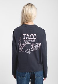 Person mit langen blonden Haaren, die ein schwarzes Langarmshirt mit einer Grafik von einem rosa Taco, scharfer Soße, Chips und Getränken auf dem Rücken trägt.
