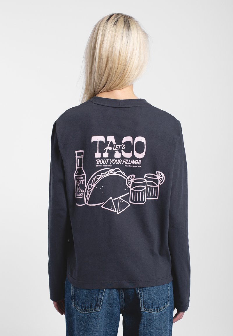 Person mit langen blonden Haaren, die ein schwarzes Langarmshirt mit einer Grafik von einem rosa Taco, scharfer Soße, Chips und Getränken auf dem Rücken trägt.