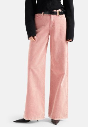 Persona che indossa pantaloni di velluto a coste rosa a vita alta e gamba larga, cintura nera con fibbia dorata, top nero aderente a maniche lunghe e scarpe nere a punta.