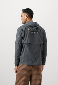 Chaqueta con capucha gris, con una abertura trasera, que presenta un diseño reflectante y puños elásticos; combinada con pantalones de cuadros marrones.