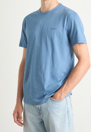 Mężczyzna w gładkim niebieskim t-shircie z krótkim rękawem z logo "BOSS" i jasnoniebieskich dżinsach, jedna ręka w kieszeni, wykadrowany w okolicy szyi i ud.
