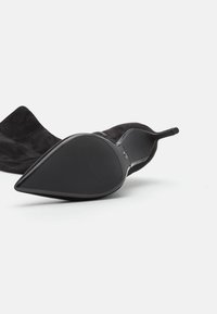 Svart mocka högklackad sko med spetsig tå, med en slät gummisula och en smal häl, sedd från sidan.