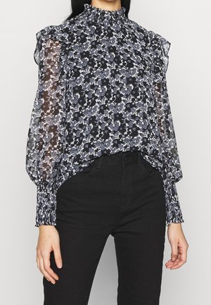 Femme portant un chemisier floral transparent à manches longues avec un col montant froufroutant et des poignets, associé à un pantalon noir taille haute.