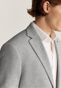 Blazer grigio chiaro in tessuto strutturato, con revers a lancia e chiusura con un solo bottone, indossato sopra una camicia bianca con collo.