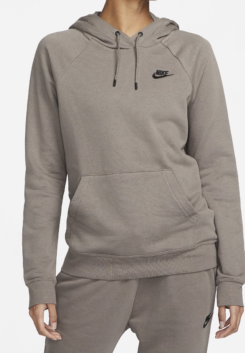 Personne portant un sweat à capuche Nike marron clair et un pantalon de survêtement assorti avec des logos Nike noirs, debout devant un fond blanc uni.