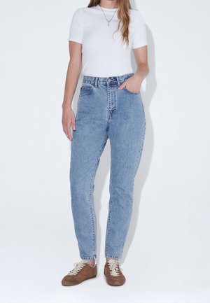 Femme portant un jean taille haute bleu clair, un haut blanc à manches courtes, des baskets marron et un collier pendentif argenté, debout devant un fond blanc.