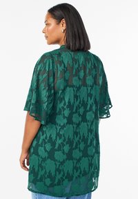 Zelená priehľadná blúzka s kvetinovou výšivkou, širokými zvonovými rukávmi a voľným strihom, v kombinácii so svetlomodrými džínsami.