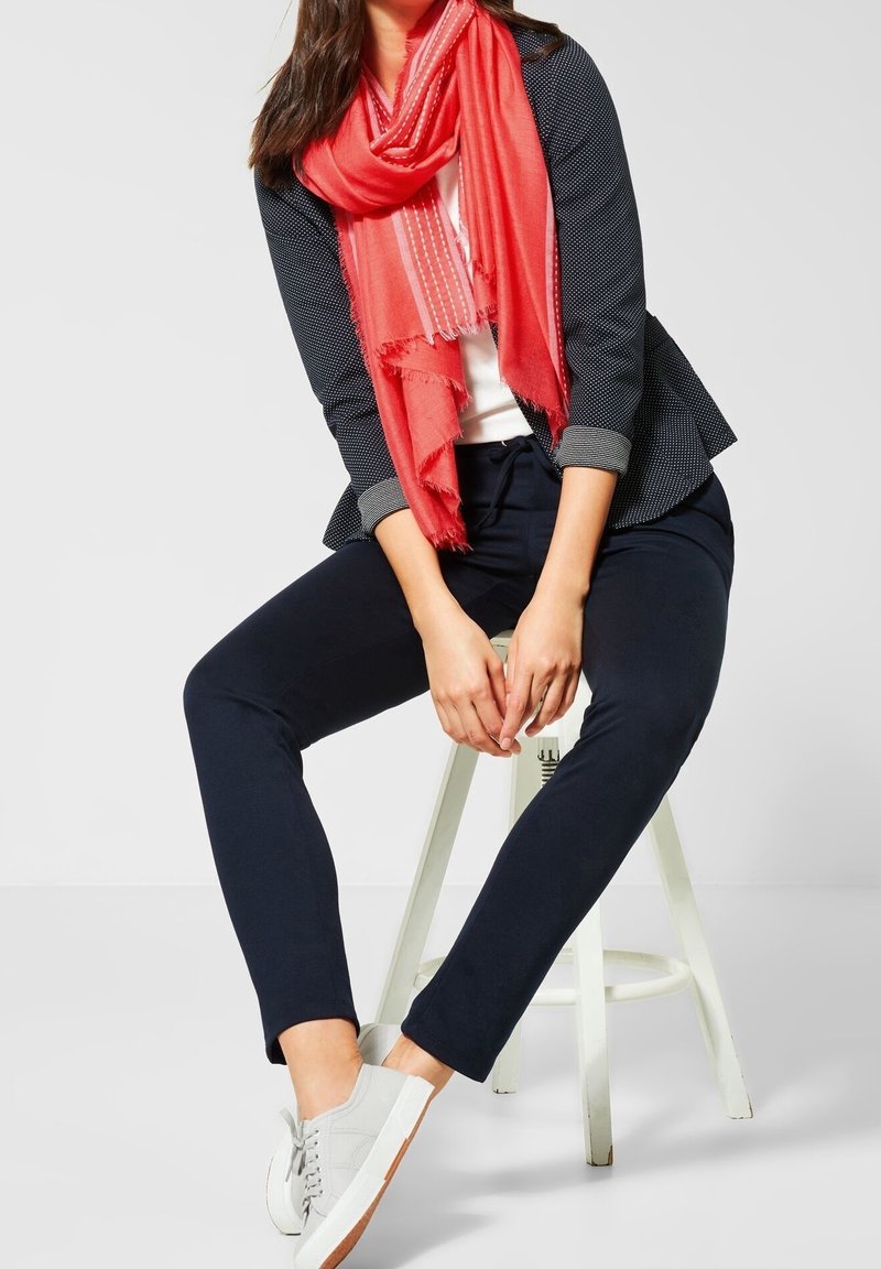 Femme assise sur un tabouret blanc portant un pantalon bleu marine, un blazer bleu marine aux manches retroussées, une écharpe corail vif, une chemise blanche et des baskets blanches.