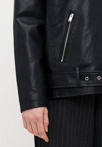 Svart syntetisk skinnjakke med sidelomme med glidelås, teksturert overflate og rett skjæring. Har en tykk kant med trykknappe-detaljer.