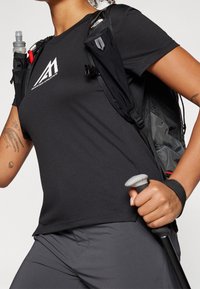 Musta urheilullinen t-paita, jossa logo, yhdistetty harmaisiin housuihin. Reppu, jossa on vesipullotaskut, ja kädessä vaellussauva.