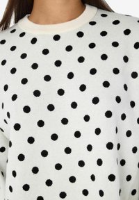 Balts džemperis ar apaļu kakla izgriezumu, kurā ir vienāda izmēra melni punkti, izgatavots no mīksta materiāla ar nedaudz teksturētu virsmu.