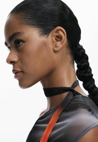 Vue de profil d'une personne avec des cheveux lisses et foncés coiffés en queue de cheval tressée. Porte un vêtement noir transparent avec une accent orange.