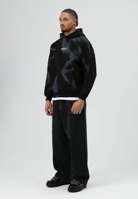 Zwarte tie-dye trui met capuchon, kangoeroezak en logo-opdruk. Gecombineerd met losse zwarte broek en dikke zwarte schoenen met gekleurde veters.