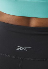 Black athletic leggings feature a gray logo at the waist, paired with a light teal sports bra, showcasing smooth fabrics and close fit.