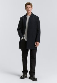 Palton din lână gri închis cu guler chinezesc, croială dreaptă, două buzunare frontale, purtat peste un pulover gri deschis și pantaloni negri.