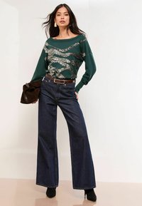 Zelená mikina so strieborným prúžkom, spárovaná s tmavomodrými širokými nohavicami a čiernymi členkovými botami. Model drží hnedú tašku.