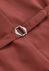 Tela de color óxido con una textura suave, que presenta un cierre de hebilla de metal plateado. El diseño incluye pliegues angulados y acentos puntiagudos.