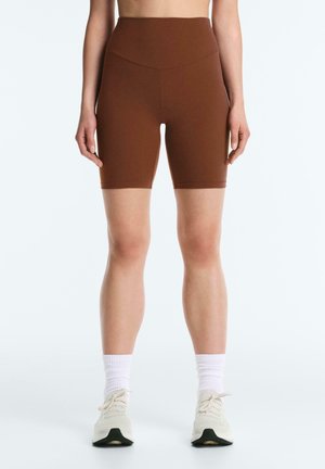 Osoba ubrana w brązowe, wysokie szorty rowerowe, białe skarpety sportowe i białe buty sportowe, stojąca na gładkim tle.