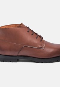 Bottes de cheville en cuir brun avec une finition lisse, un design à lacets et un intérieur texturé couleur beige. Équipées d'une robustes semelle en caoutchouc noir.