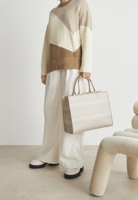 Osoba v béžovém a krémovém svetru a širokých bílých kalhotách drží velkou béžovou vzorovanou tašku a stojí vedle moderní béžové židle.