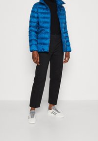 Blå puffjacka med horisontell quiltad textur, dragkedja, svart polotröja, svarta byxor och vita sneakers med gråa ränder.