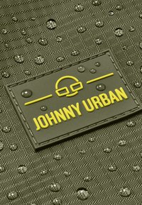 Voděodolná, strukturovaná šedá tkanina s žlutou nášivkou "JOHNNY URBAN", která má vzvednuté detaily a kapky vody na povrchu.