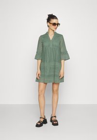 Rochie verde din dantelă cu decolteu în V și mâneci evazate trei sferturi, prezentând modele geometrice și un fustă evazată; asortată cu sandale negre.