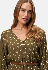 Olivengrünes Kleid mit V-Ausschnitt, floralem Druck in Weiß und Gelb, Knopfdetails und einem braunen Gürtel, der die Taille betont. Lange Ärmel.