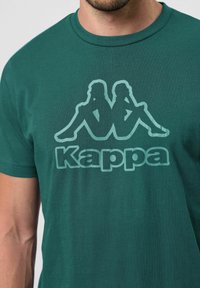 Zelené bavlněné tričko s velkým, vybledlým logem a dvěma sedícími postavami nad textem "Kappa" ve vyznačeném tučném písmu.